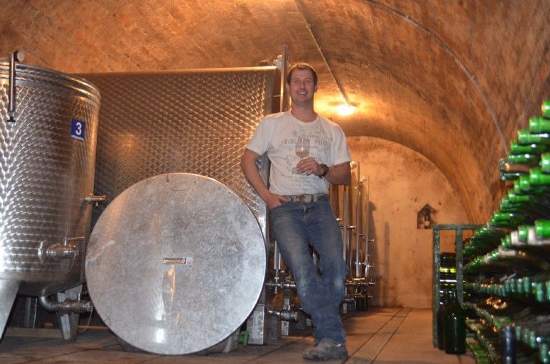 A man stands in a wine cellar next to large stainless steel tanks and bottles of wine.