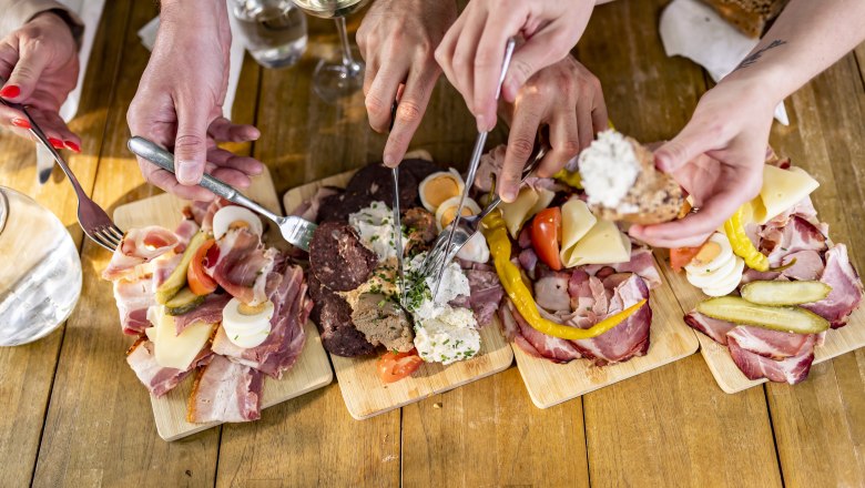 Several hands reach for a selection of sausage, cheese and bread on wooden platters.