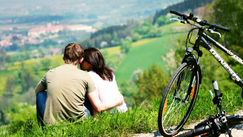 A couple is sitting on a meadow with a view of the countryside, with bicycles parked next to them.