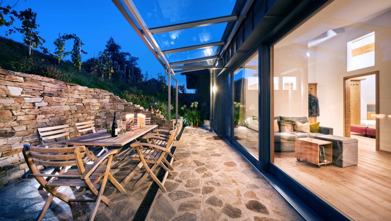 Terrace with wooden furniture and glass roof, view into the illuminated living room.