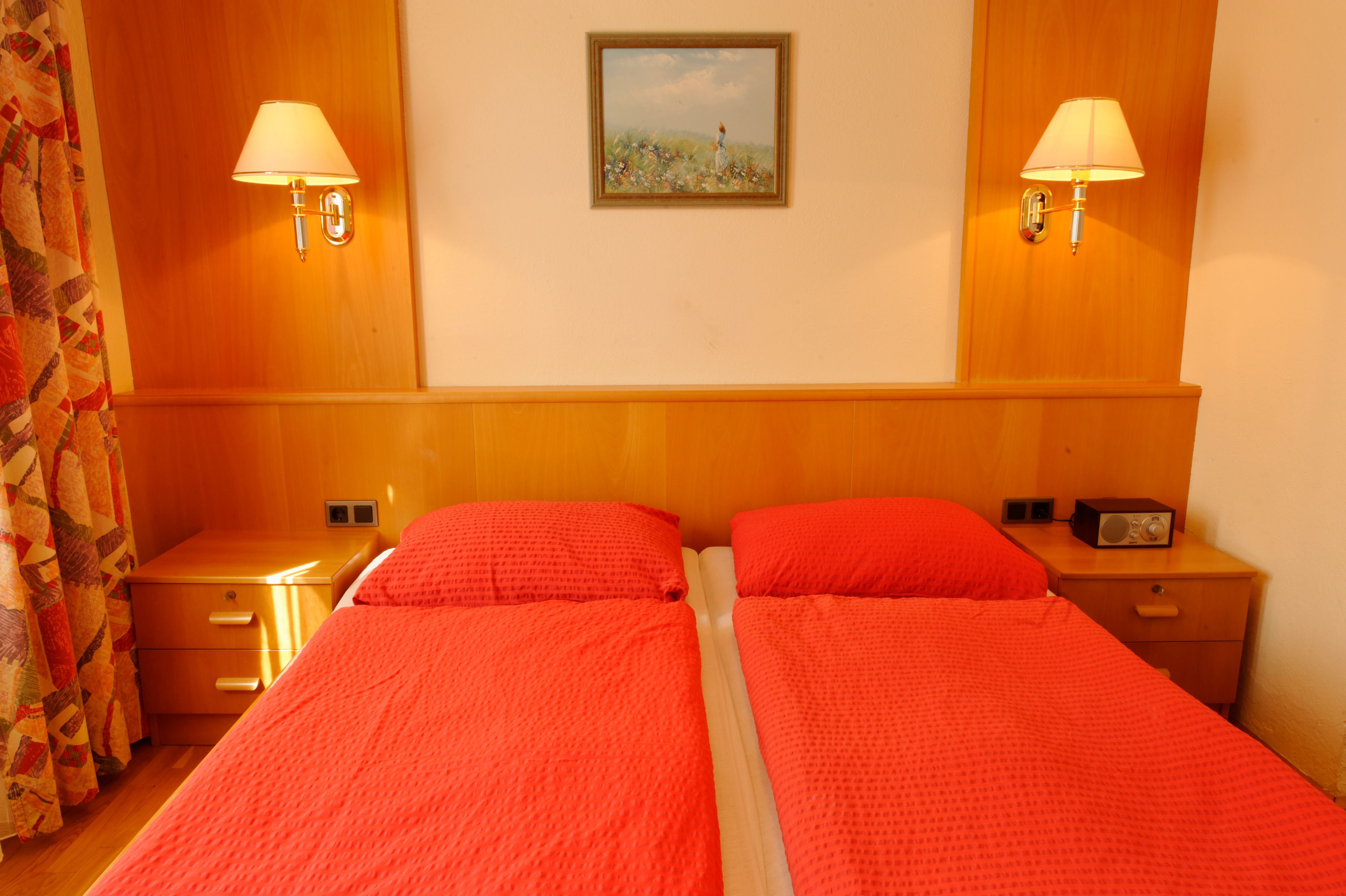 Guest room with two single beds, red comforters, wooden furniture and wall lamps.
