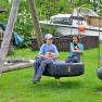 Four children sit on a swing in the garden and hold animals. There is a boat in the background.