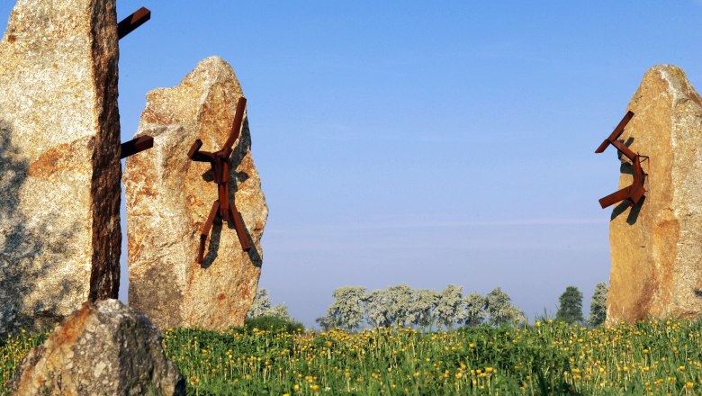Stone sculptures with metal figures on a meadow under a blue sky.