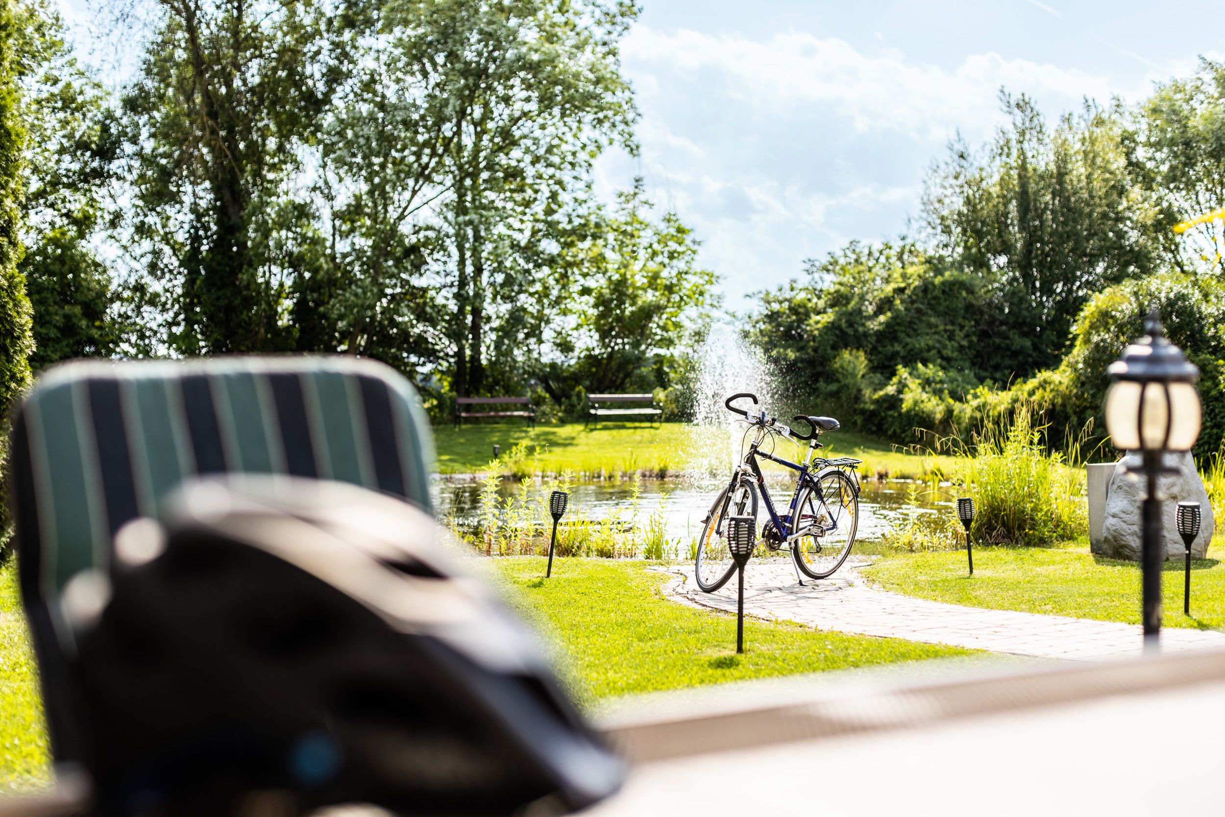 A bicycle is parked next to a pond with a fountain in a sunny garden.