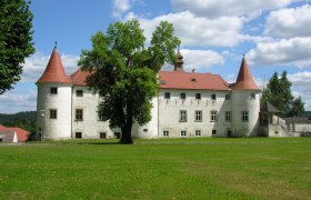 A historic castle with red roofs and towers, surrounded by green meadows and trees, under a blue sky with white clouds.