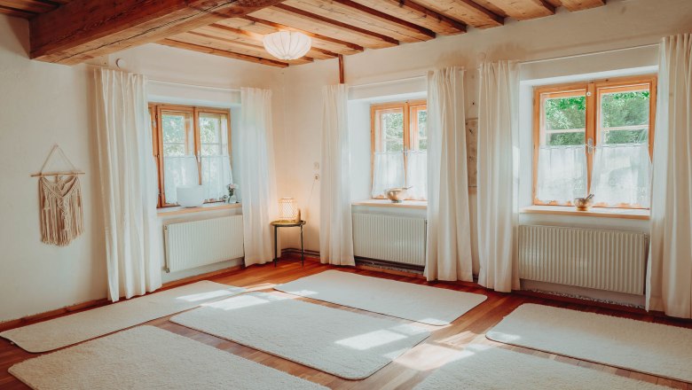 A bright yoga room with a wooden floor, white mats and large windows with curtains.