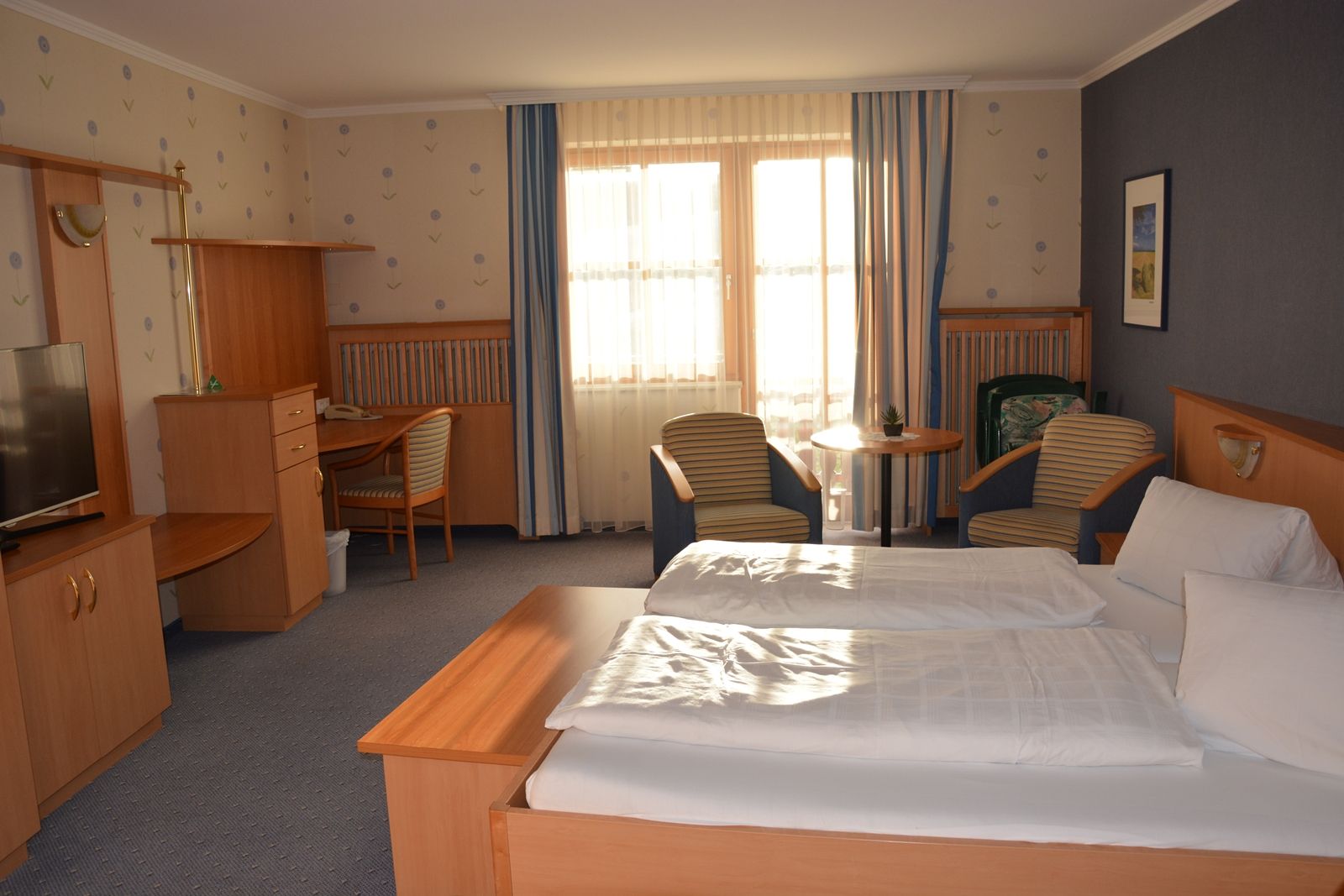 A cozy hotel room with a double bed, desk, TV and two armchairs in front of a window with curtains.