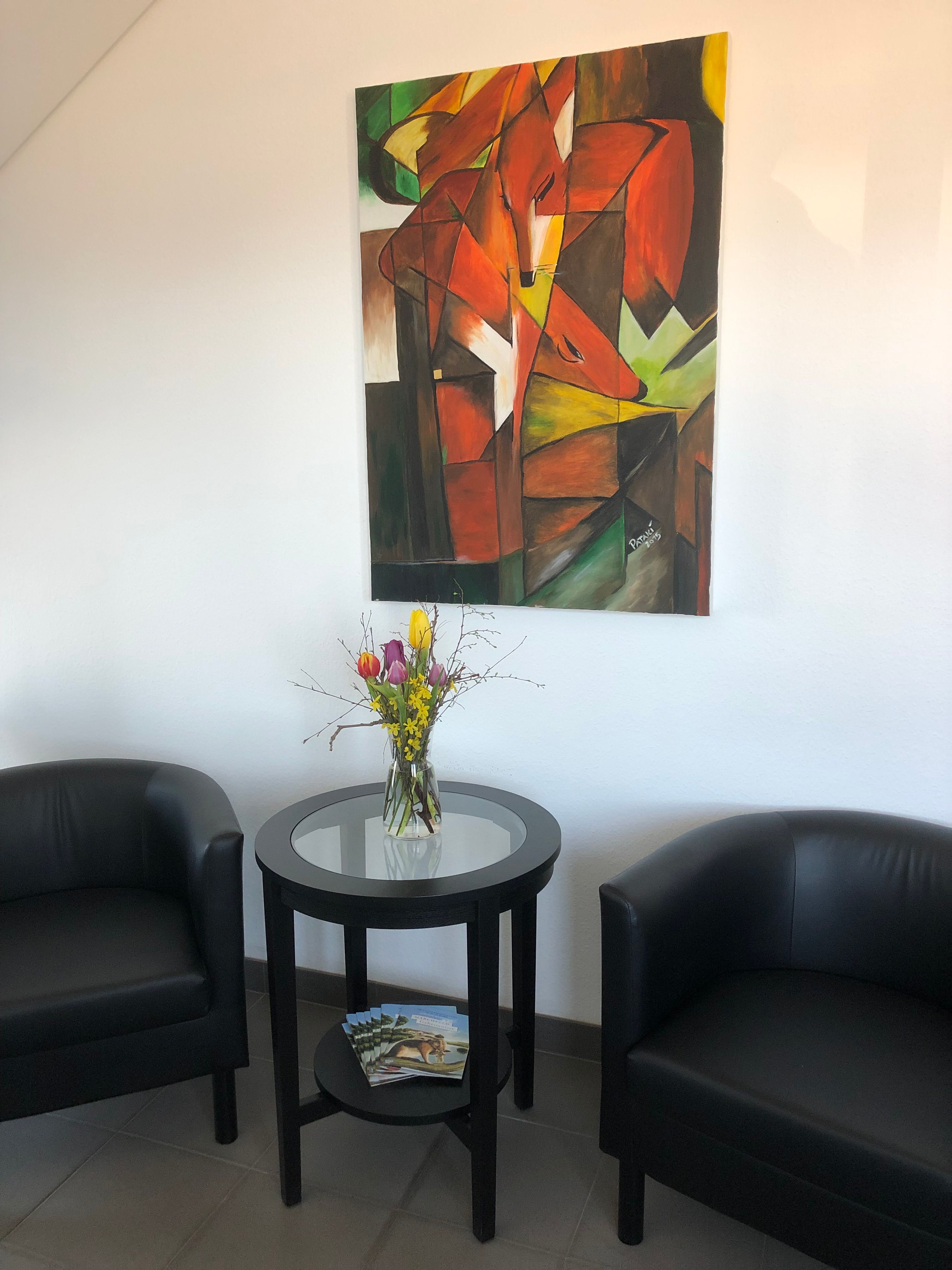 Two black armchairs and a round table with flowers in a room. An abstract painting with a fox motif hangs on the wall.
