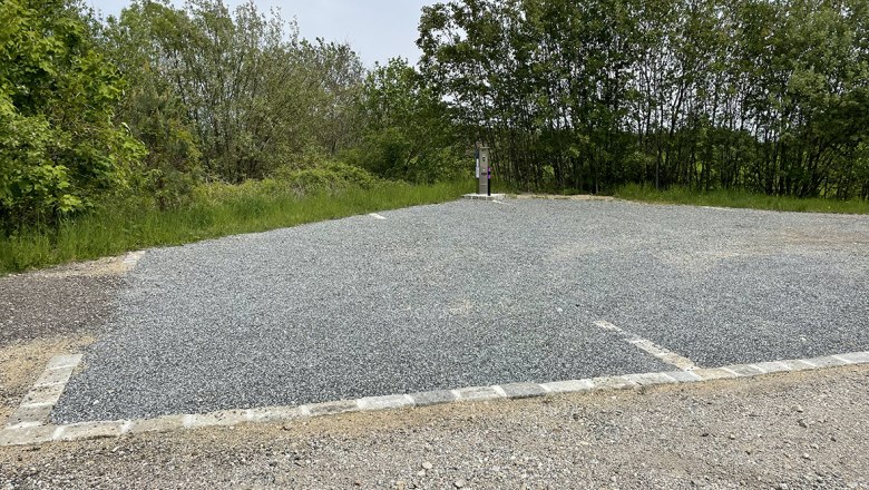 An empty camper van site with a gravel floor, surrounded by trees and a lantern.