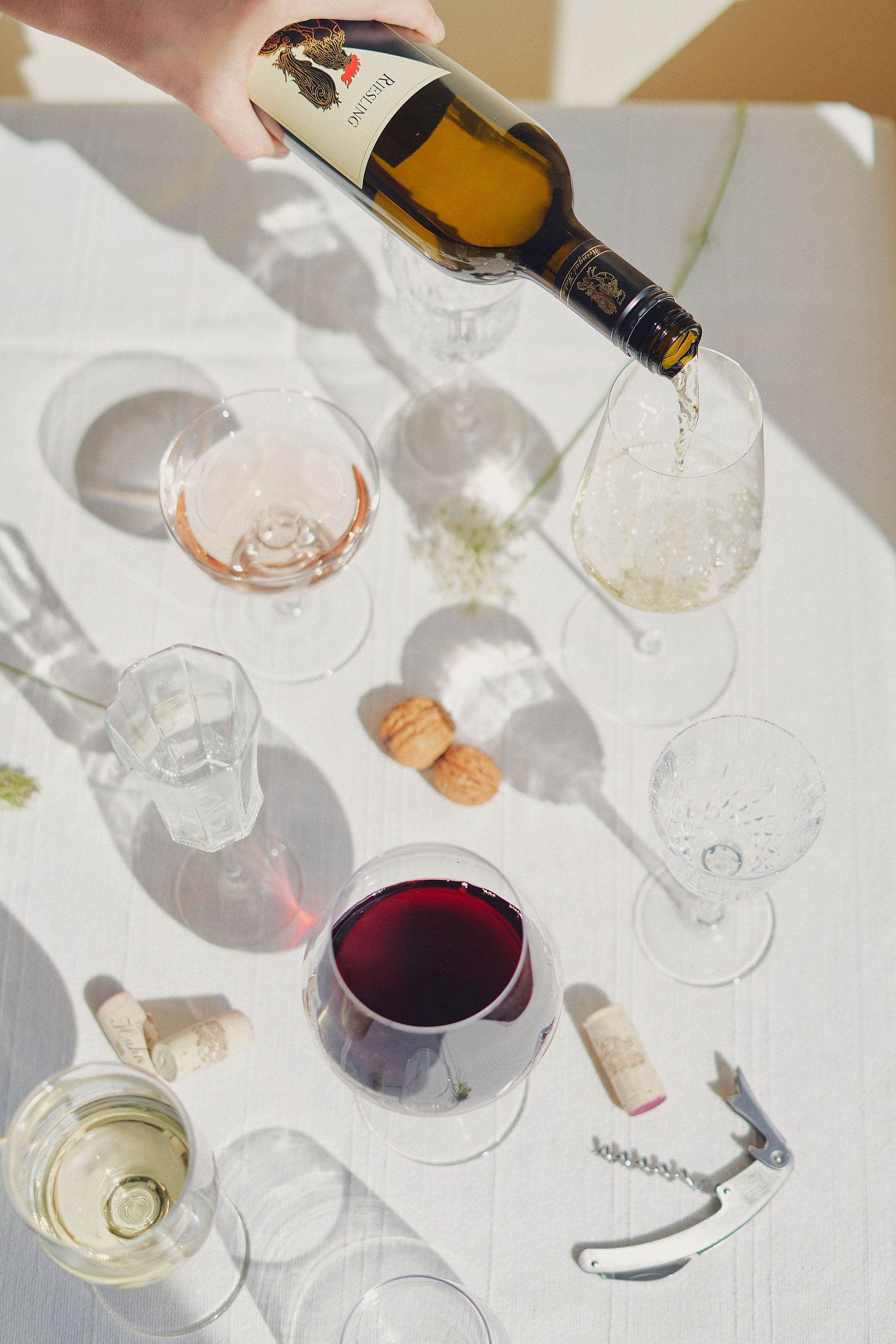 Wine bottle is poured into a glass on a table with various wine glasses and corks.