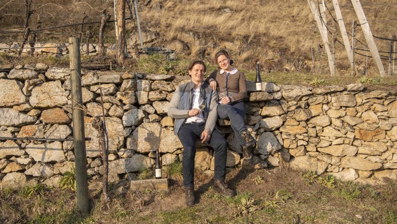 Two people are sitting on a stone wall in a vineyard holding wine glasses.
