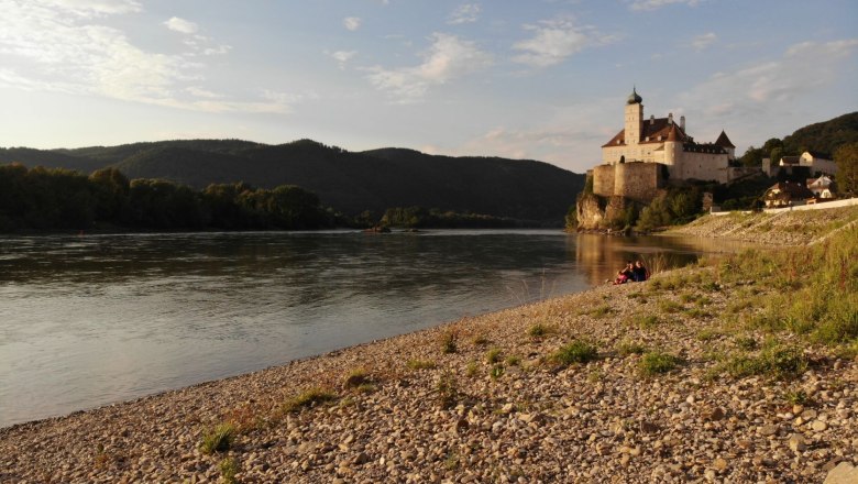 Pebble beach on the Danube with castle in the background at sunset.