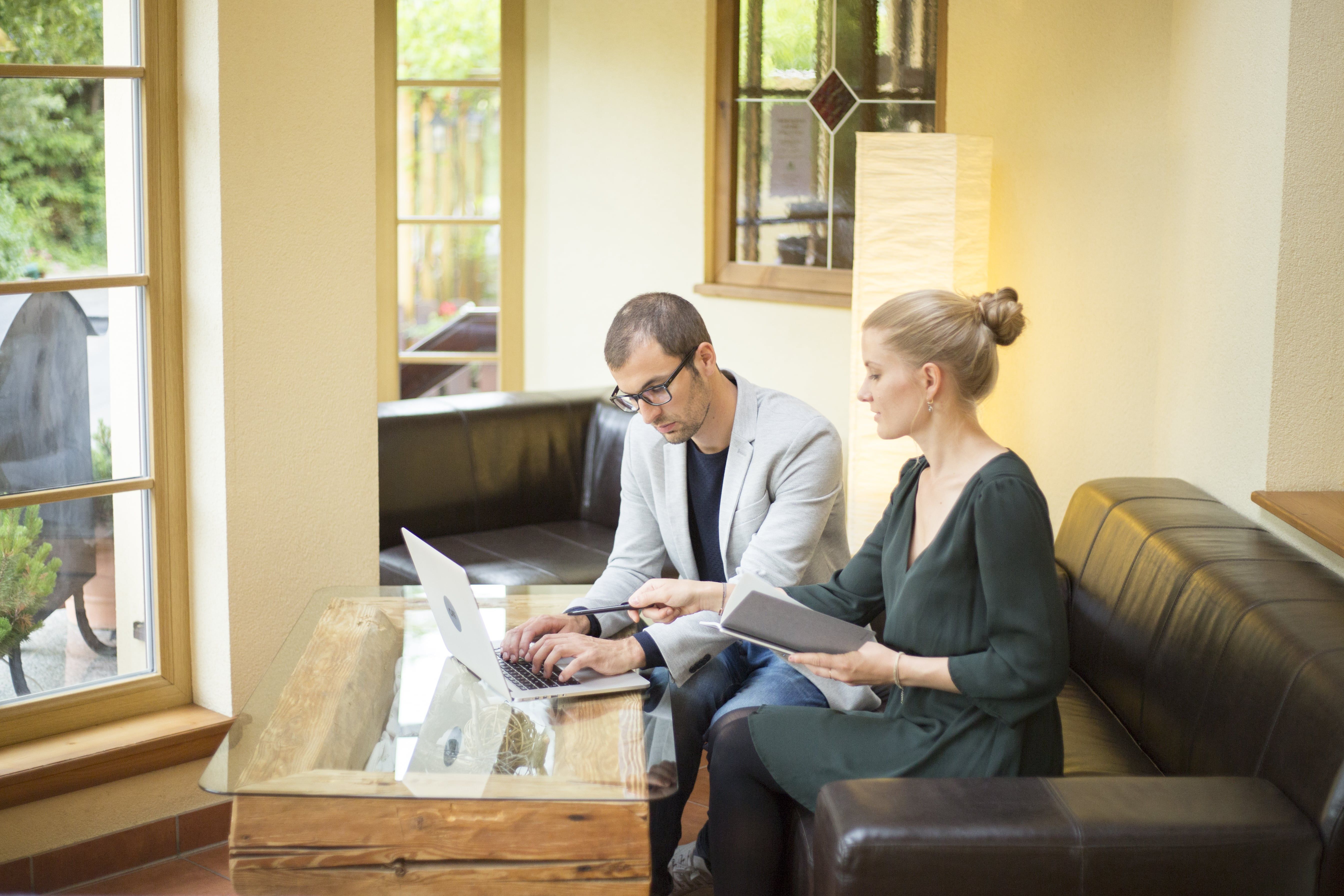 Two people work on a laptop in a cozy room with large windows.