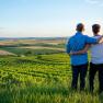 A couple stands embracing on a hill overlooking a vast wine landscape at sunset.