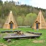 Two wooden tepees on a meadow in front of a forest.