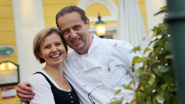 A smiling couple in traditional dress in front of a yellow building.