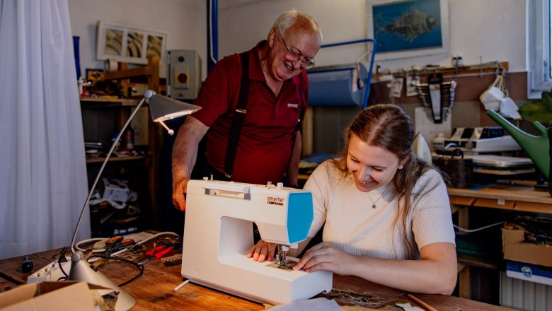 A young woman sews on a sewing machine while an older man looks on with a smile. The room is equipped with tools and materials.