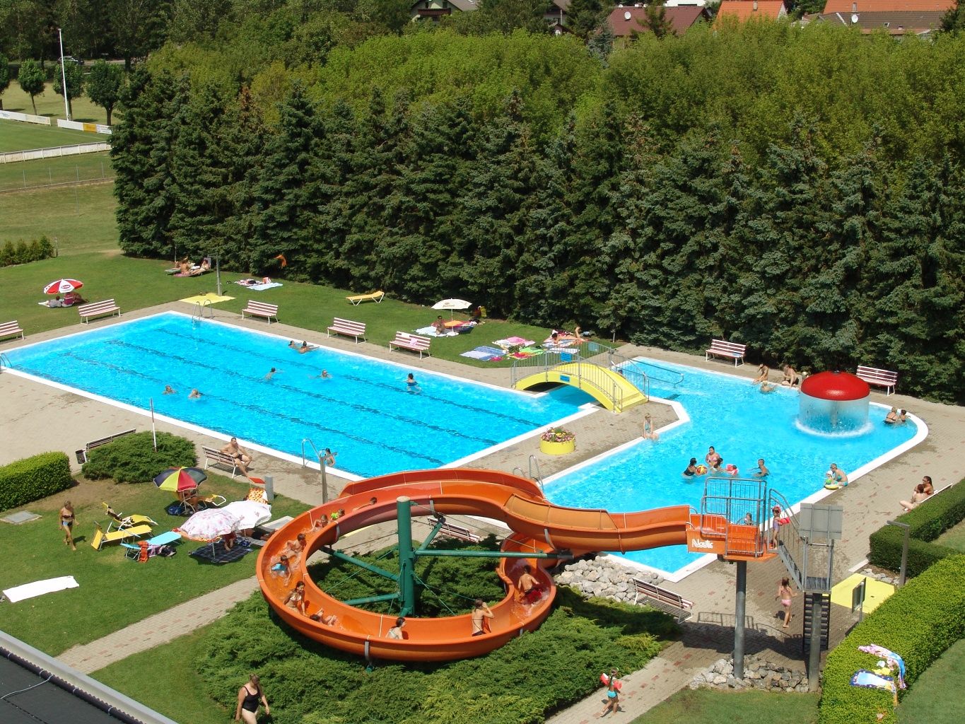 Aerial view of an outdoor pool with slide, two pools and sunbathing area.