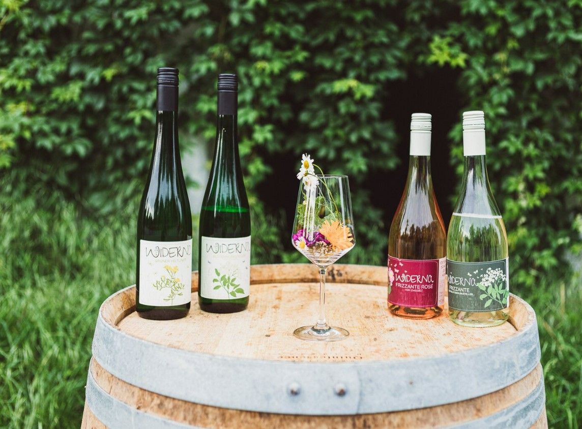 Four wine bottles and a wine glass with flowers on a wooden barrel in front of a green hedge.