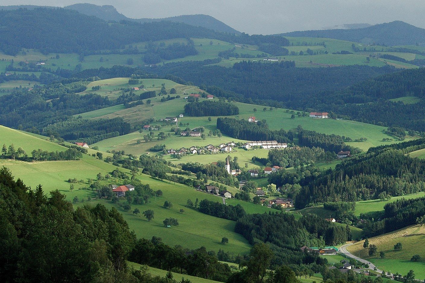 Panoramic view of a green, hilly landscape with scattered houses and forests.