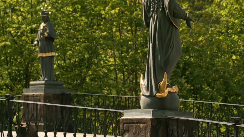 Two statues on a bridge with railings, surrounded by green trees.