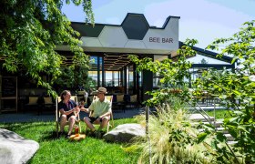 Two people sit in the garden in front of the Bee Bar and toast with drinks.