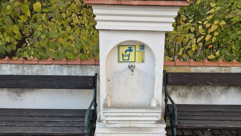 A small fountain with a tiled roof and tap, flanked by two wooden benches on a paved square.