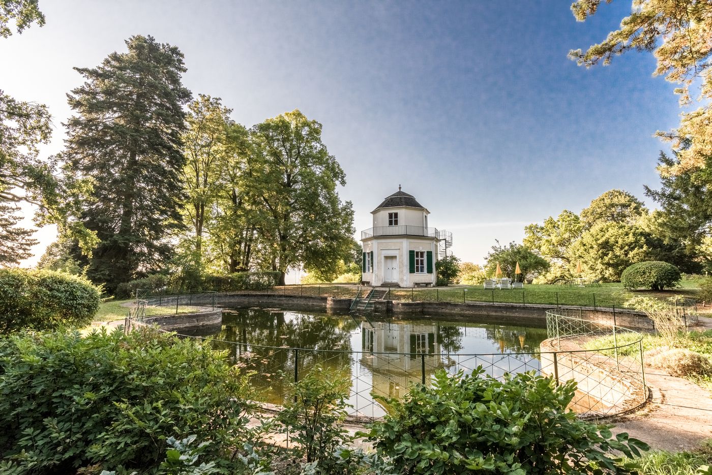 An octagonal pavilion stands next to a pond, surrounded by trees and a garden.