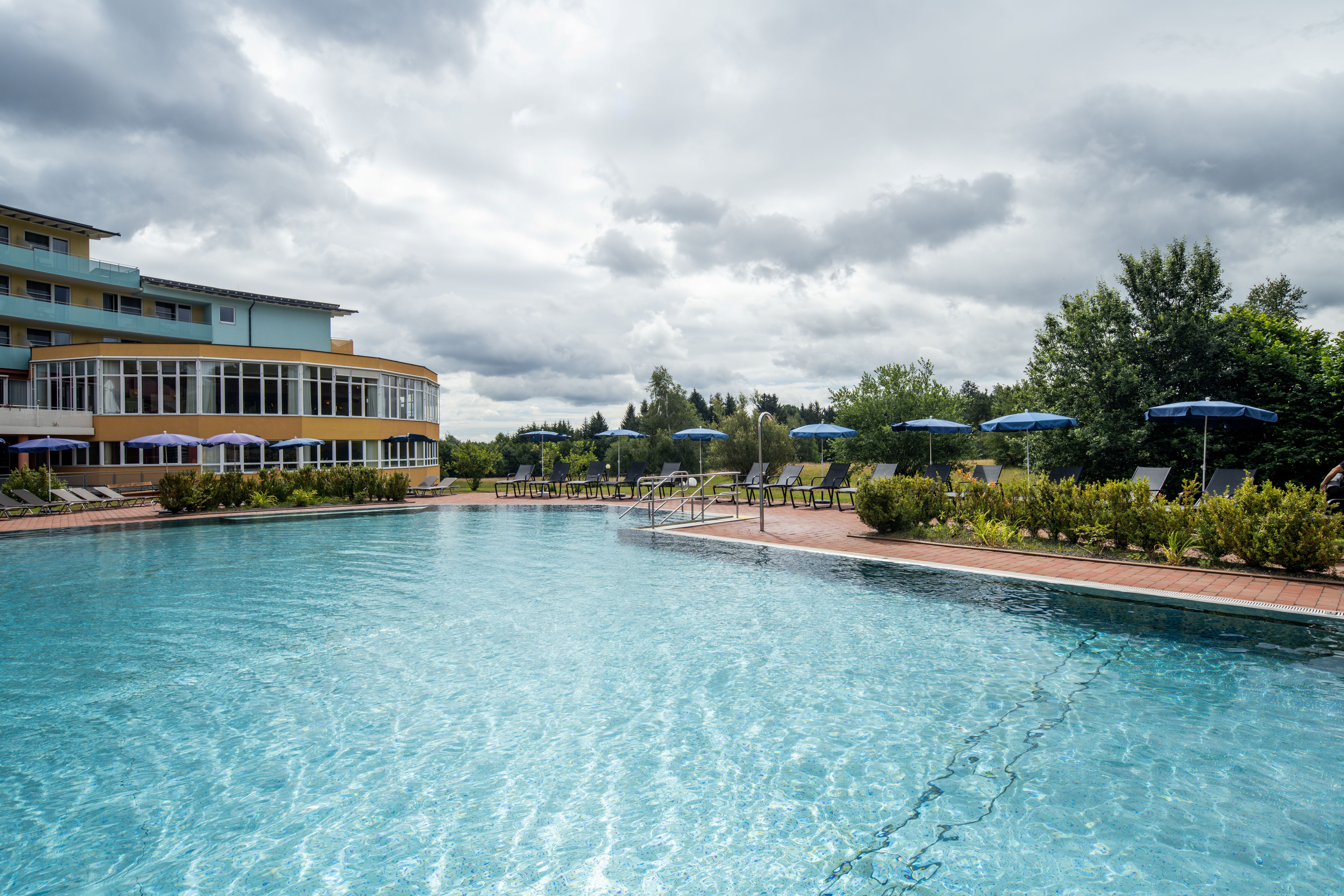 An outdoor pool with clear water in front of a building with large windows and several stories. There are lounge chairs and umbrellas by the pool.