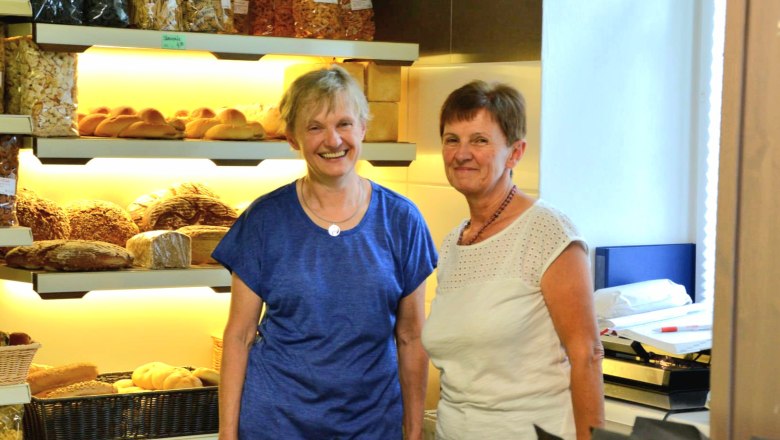 Two women in a bakery in front of shelves of bread and pastries.