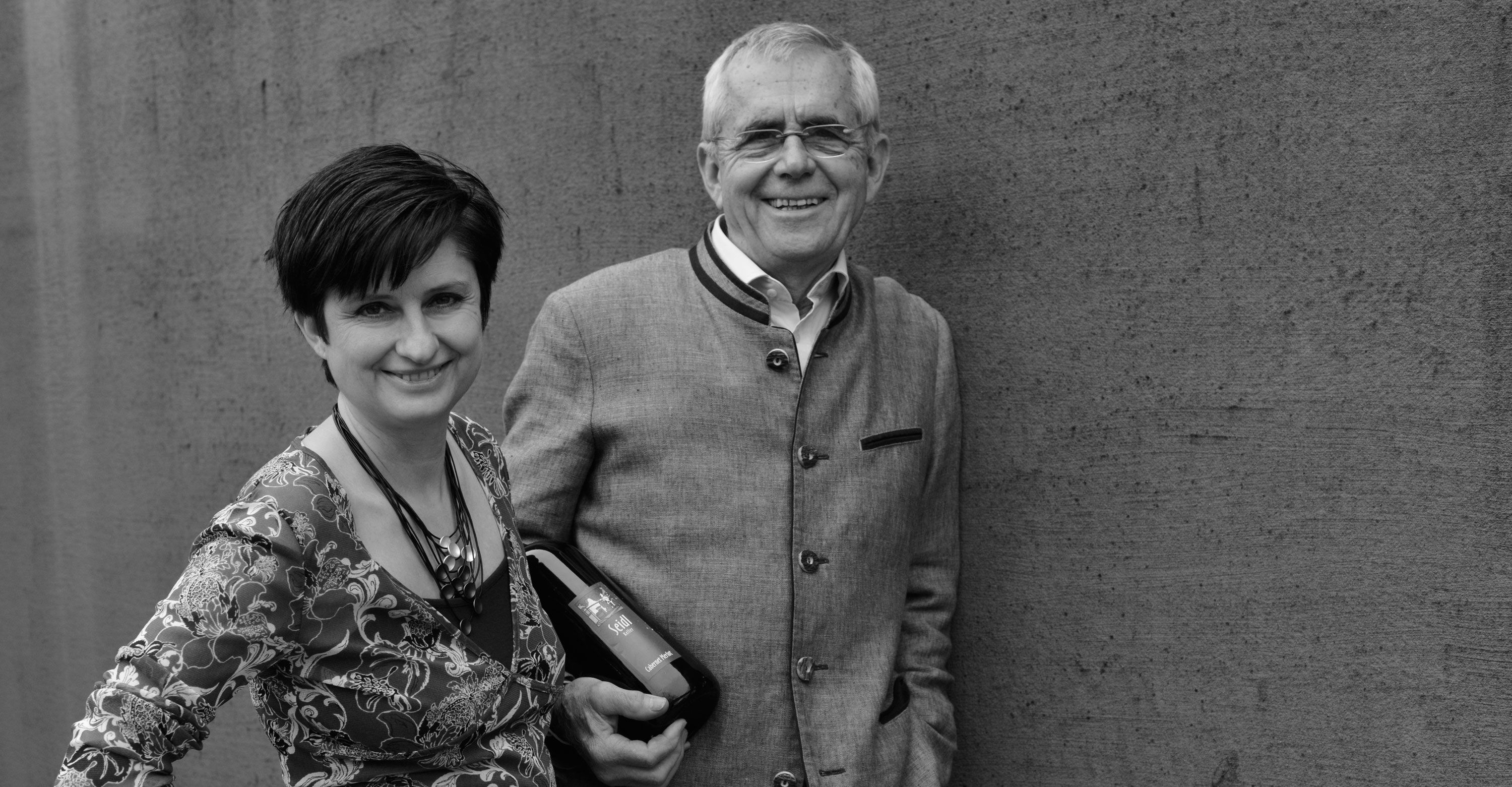 A man and a woman stand smiling in front of a wall. The man is holding a bottle of wine.