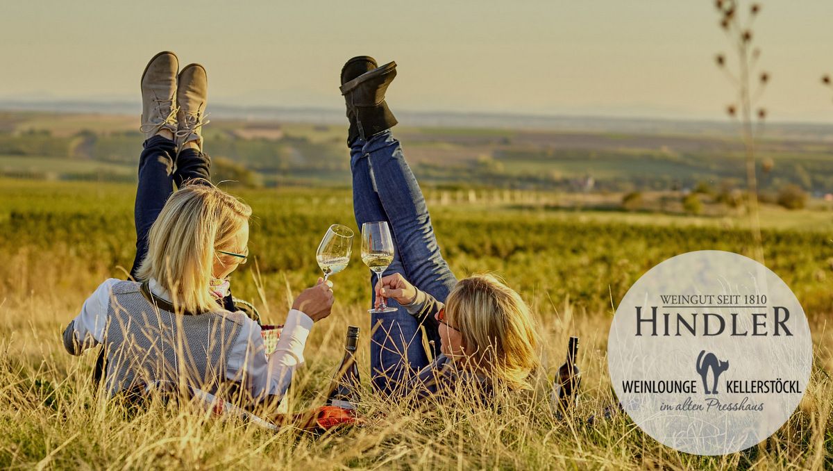 Two people are lying in a vineyard, raising their feet and holding wine glasses. The Hindler winery logo is shown.