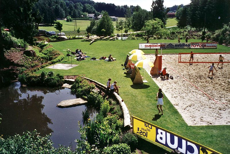 Green meadow with swimming pond, volleyball court and people relaxing and playing.