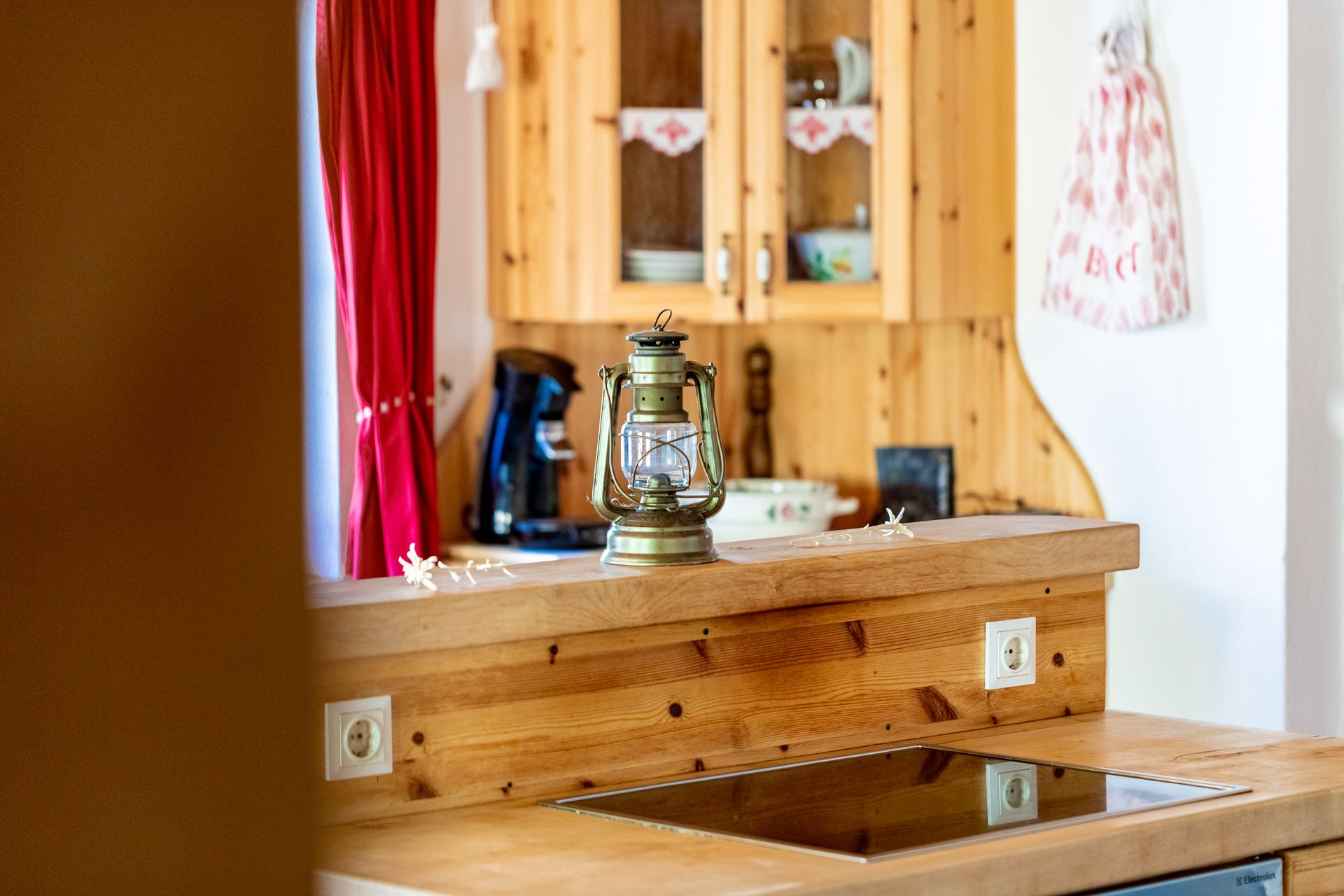 Wooden kitchen sideboard with ceramic hob, lantern on the counter, box and red curtain in the background.