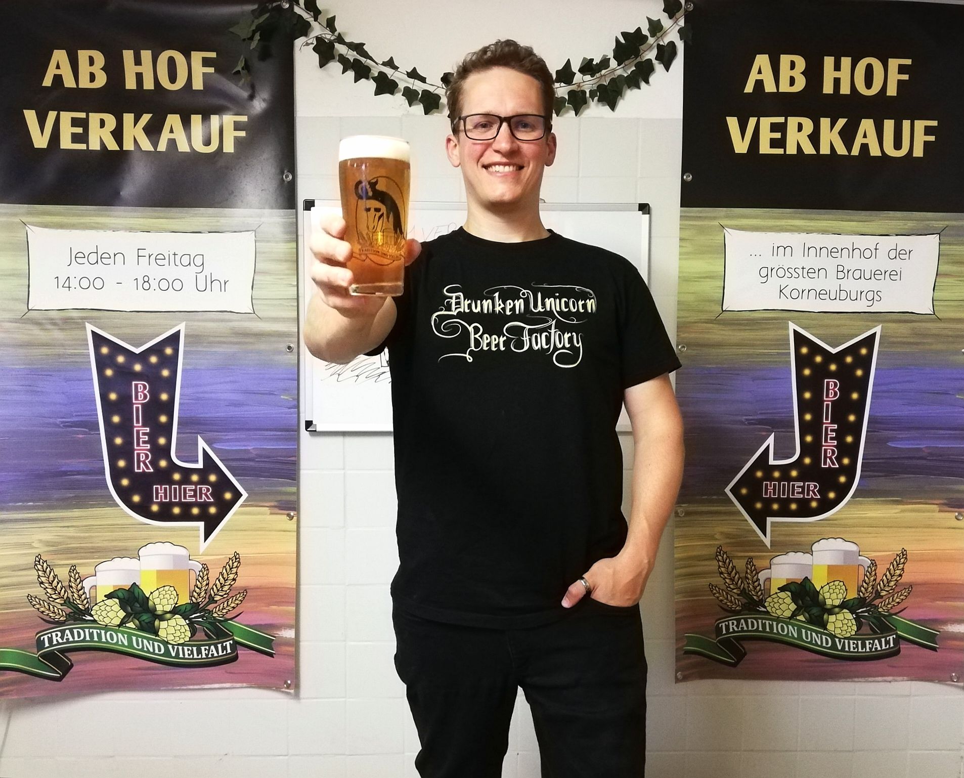 Person holding a beer glass in front of two banners with the inscription 'Ab Hof Verkauf'.