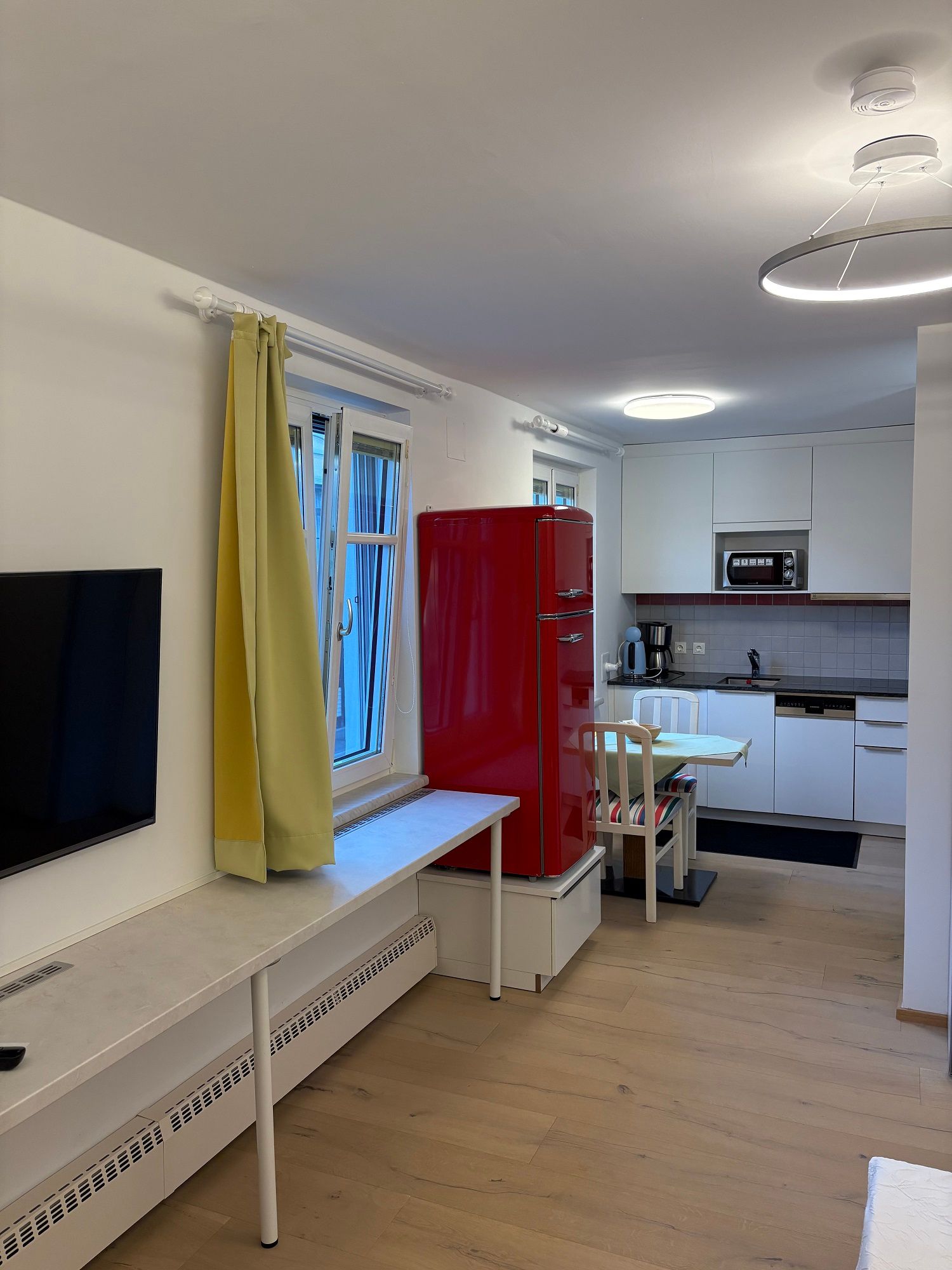 Modern living room with open kitchen, red fridge, yellow curtains and wooden floor.