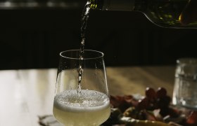 A glass of white wine is poured on a table, with a plate of snacks in the background.