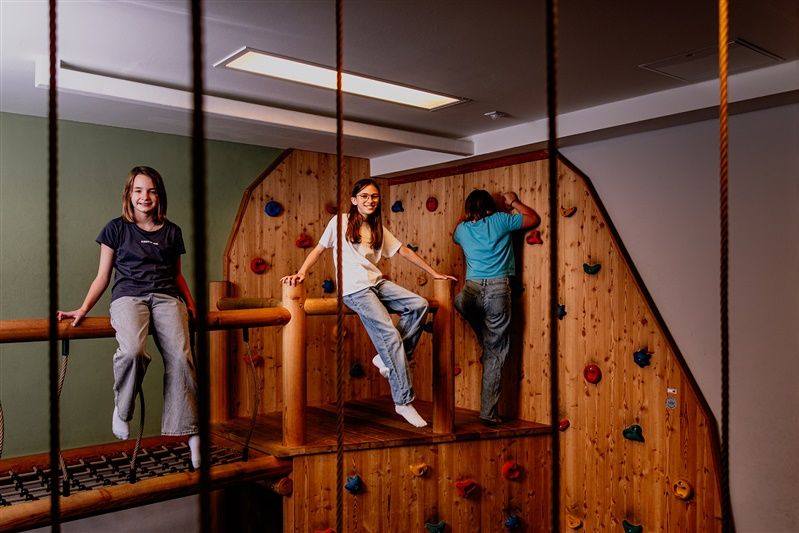 Children play in an indoor climbing area with wooden walls and colorful climbing holds.