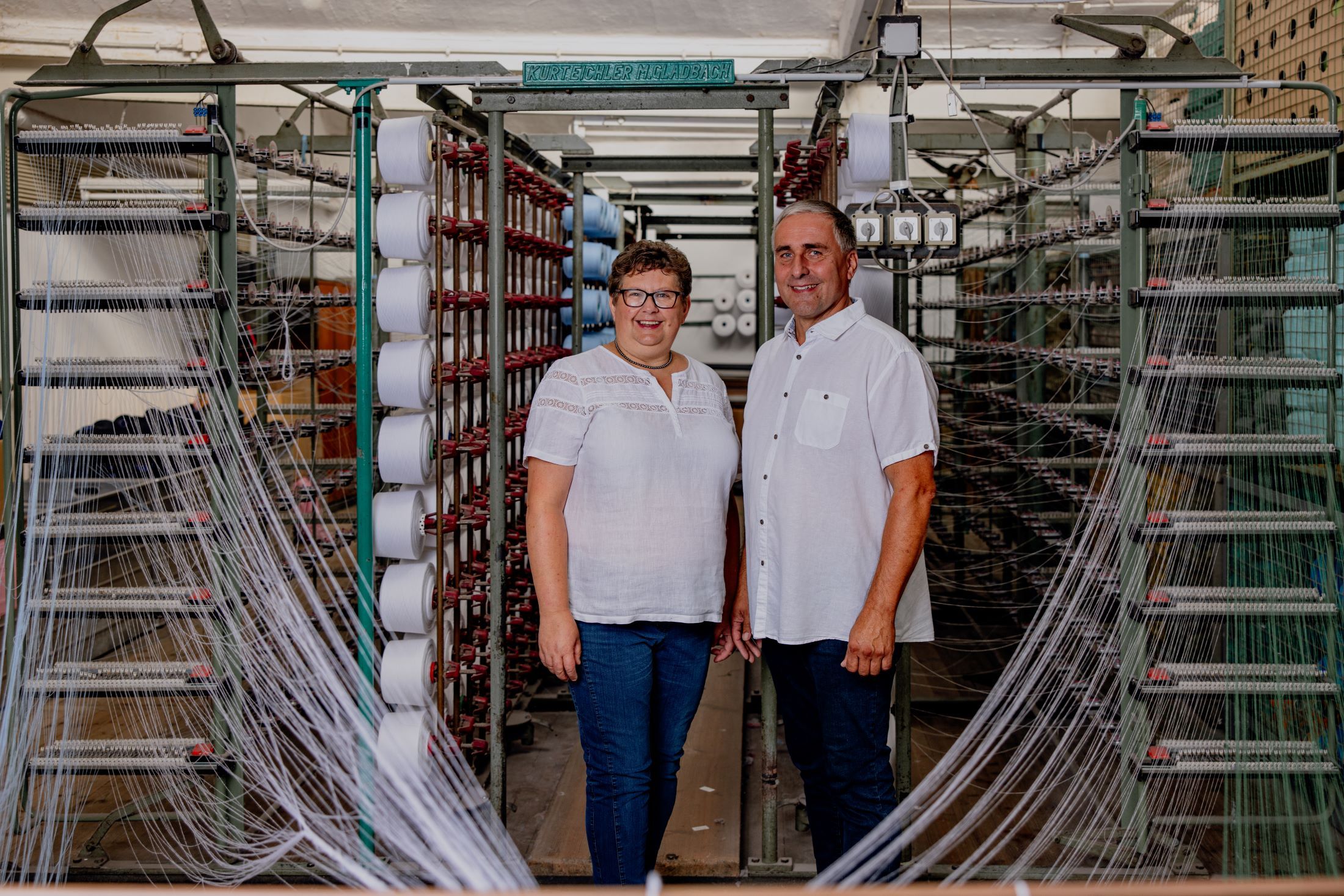 Two people are standing in front of machines with spools of thread in a textile factory.