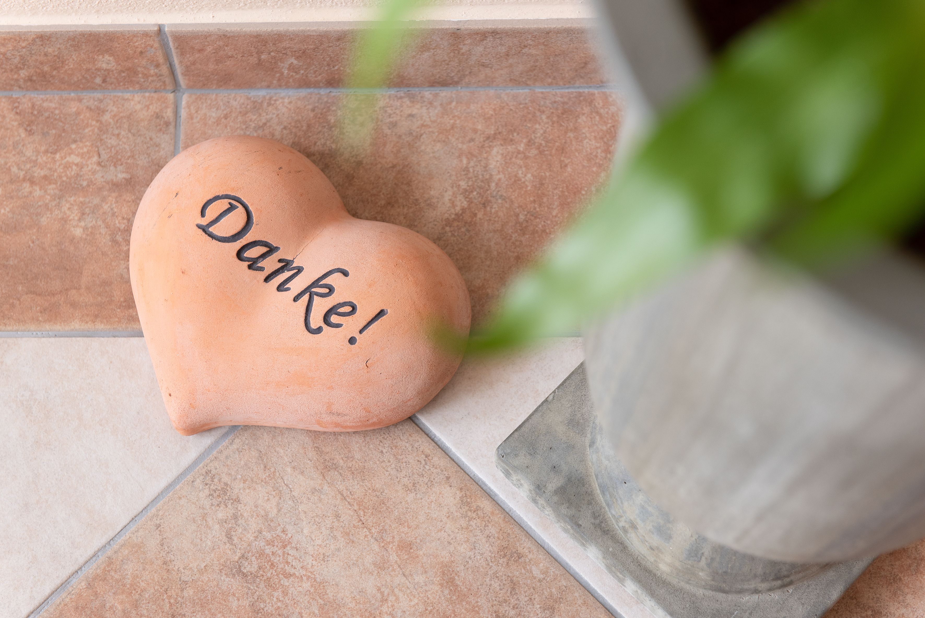 Heart-shaped stone with the inscription 'Thank you' on tiled floor next to plant pot.