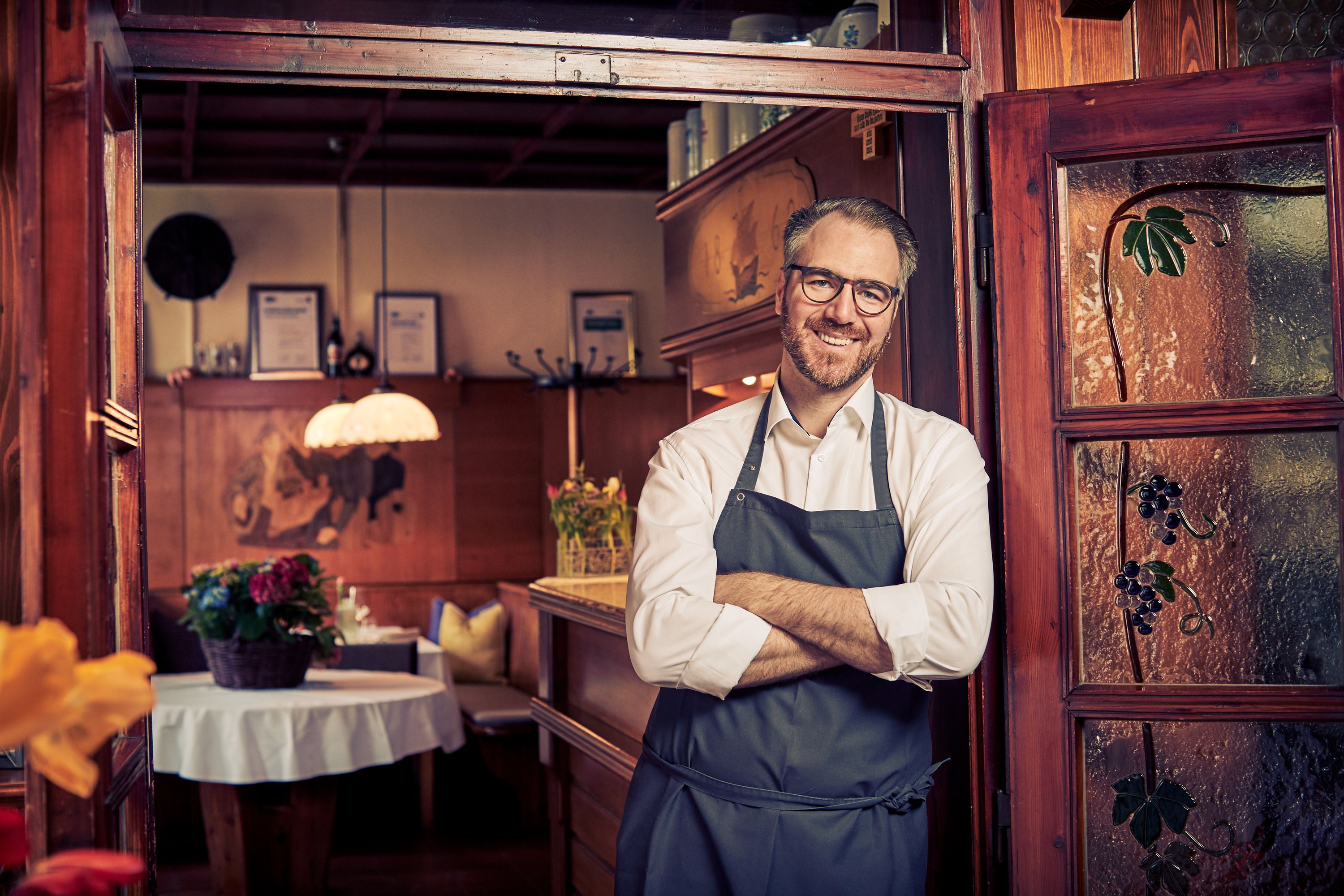 Smiling man in an apron stands in the doorway of a cozy restaurant.