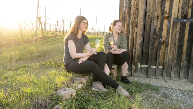 Two women sit with wine glasses in front of a wooden hut in the vineyard.