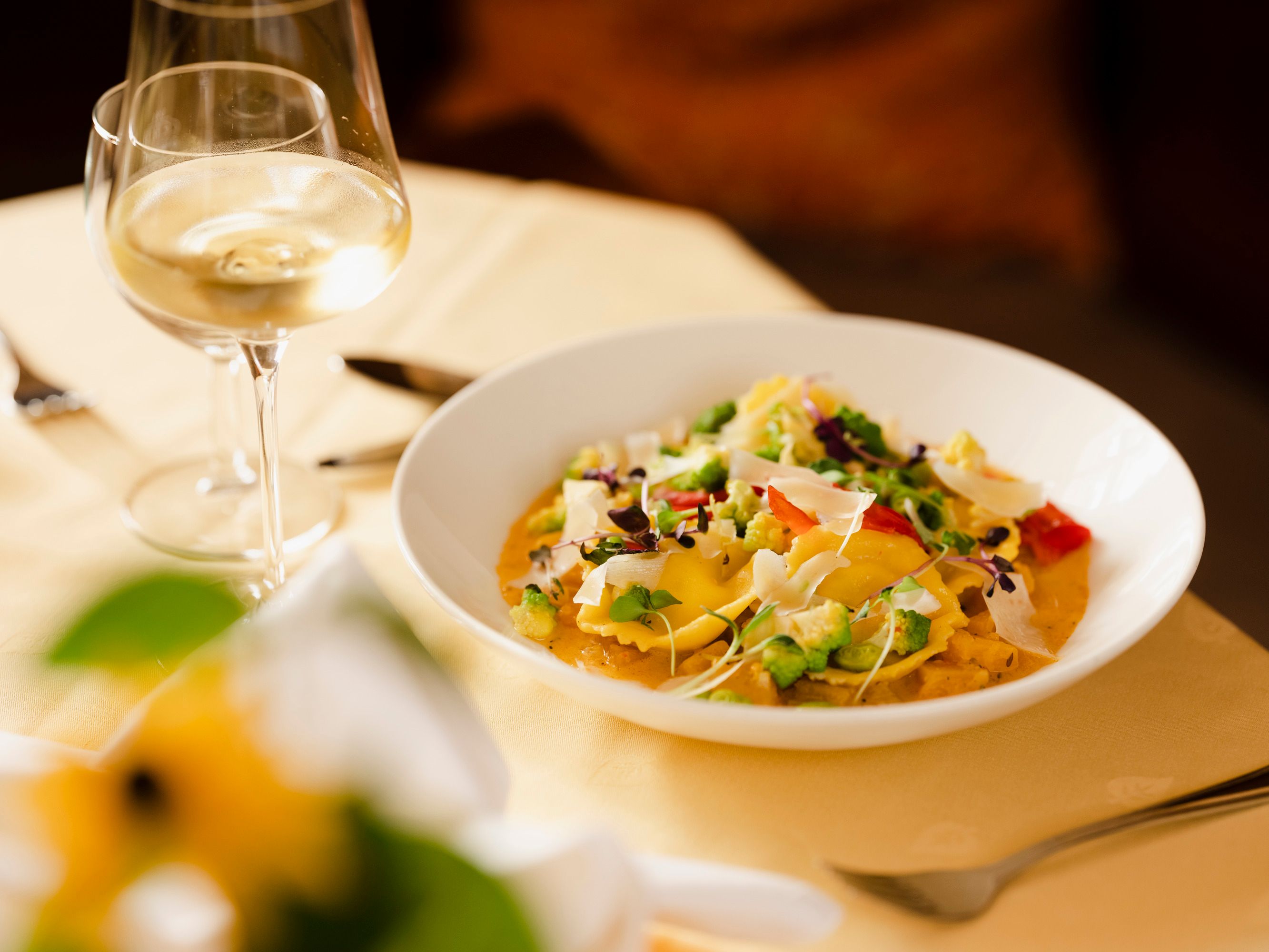 A plate of cream cheese ravioli, garnished with vegetables and herbs, next to a glass of white wine on a laid table.