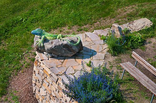A colorful lizard statue on a stone pedestal next to a bench and blue flowers.