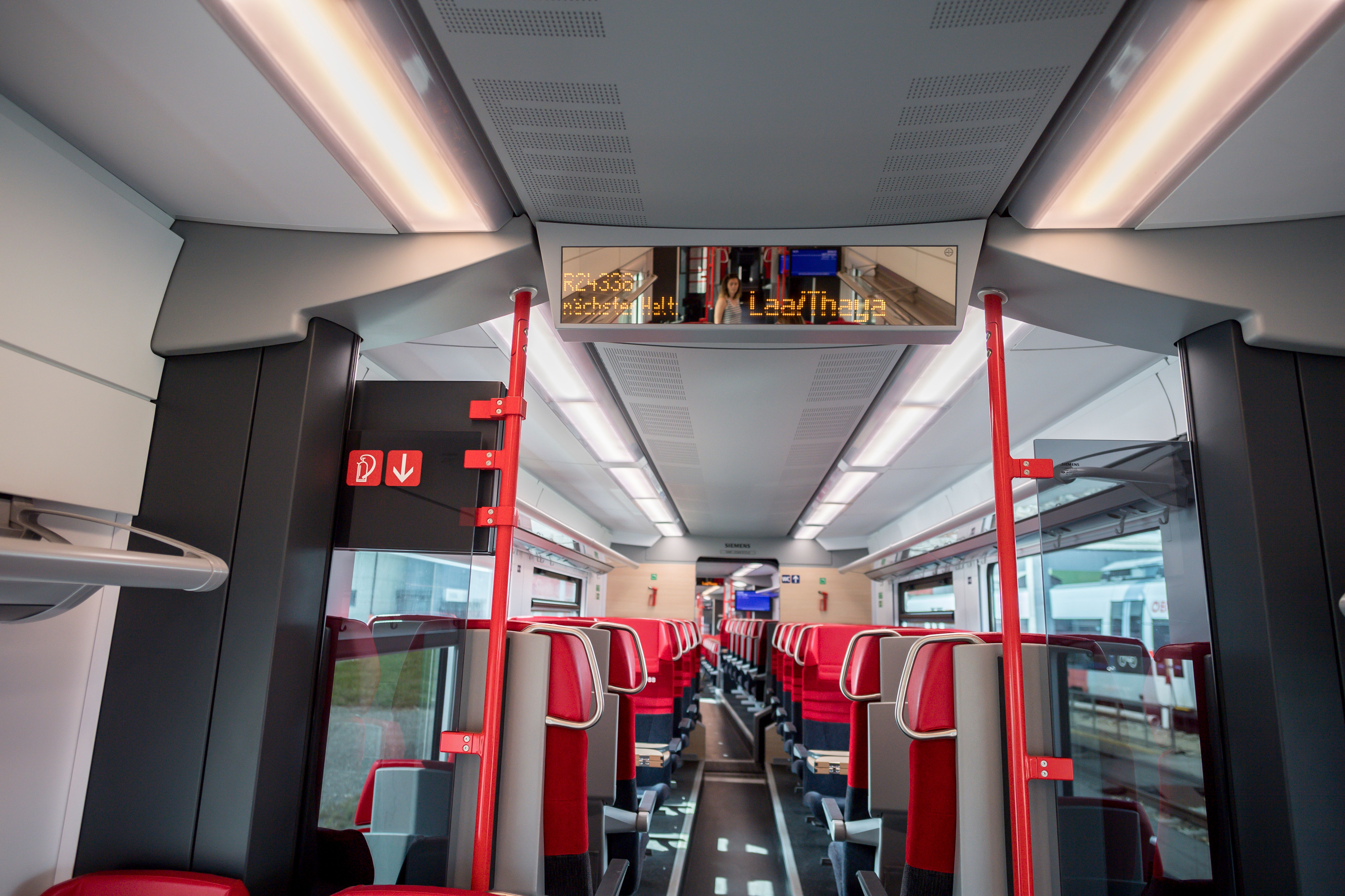 Interior view of a modern train compartment with red and gray seats and a digital display board.