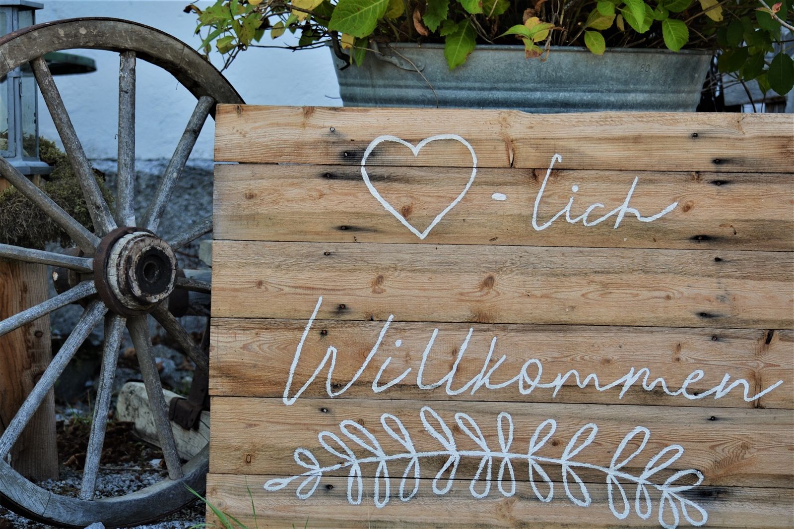 Wooden sign with the inscription 'Welcome' and a heart symbol, next to it an old wagon wheel.