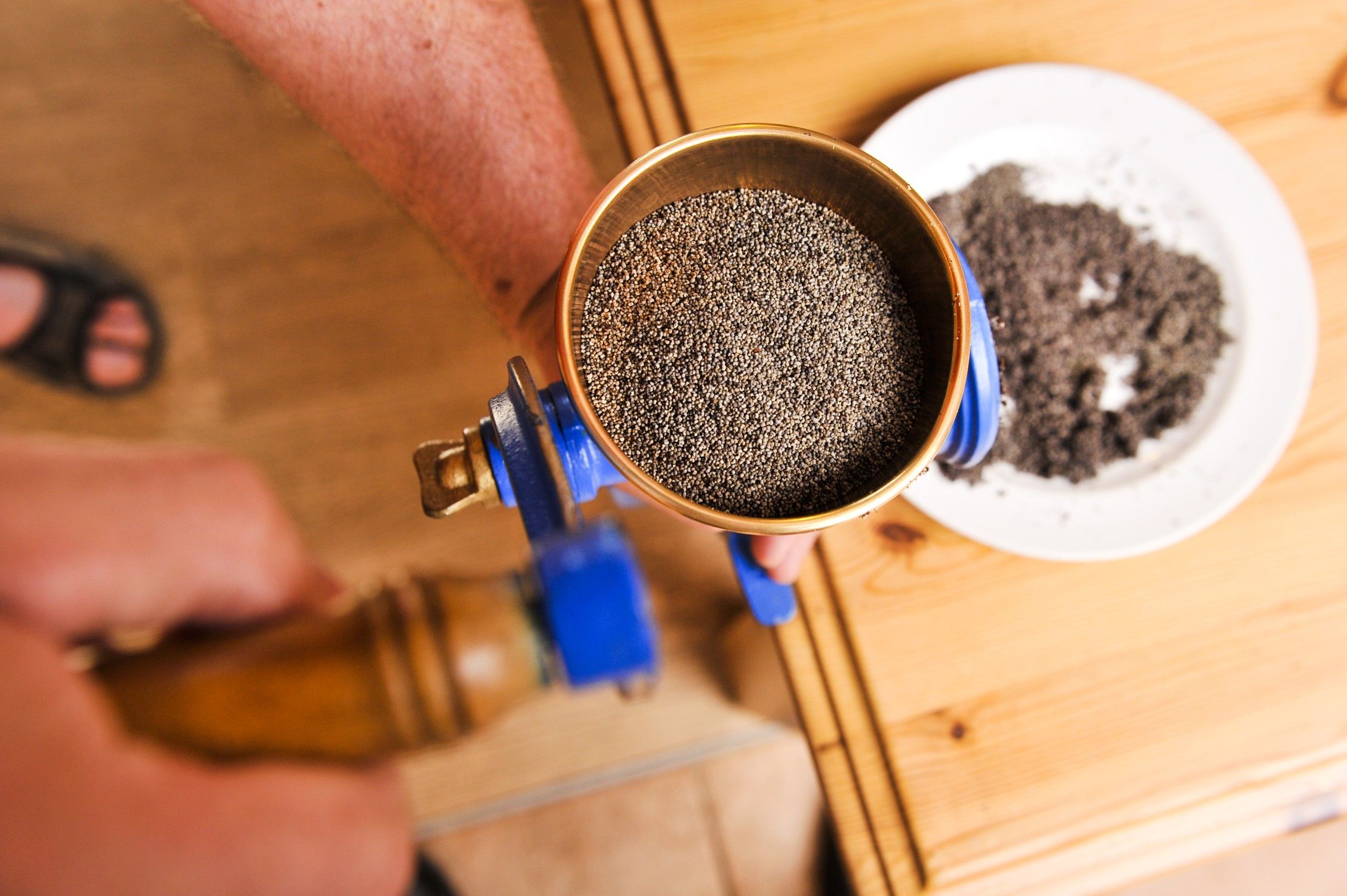 Close-up of a hand operating a mill with poppy seeds, next to it a plate with ground poppy seeds.