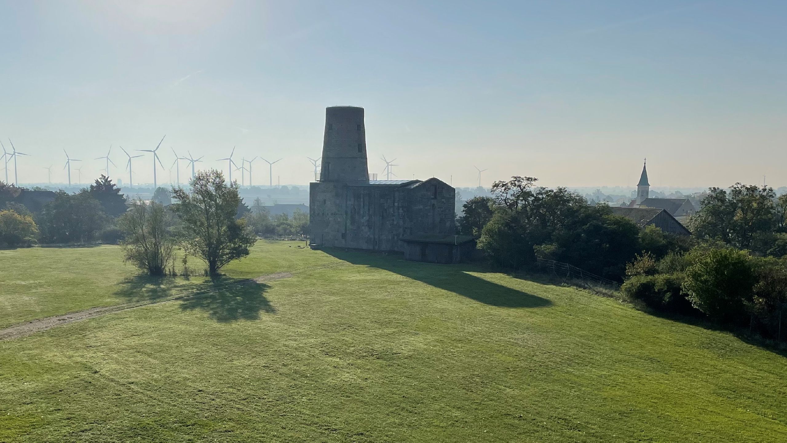 View of the Ruins: A vast green meadow with an old ruin in the center, wind turbines in the background, and a church spire to the right.