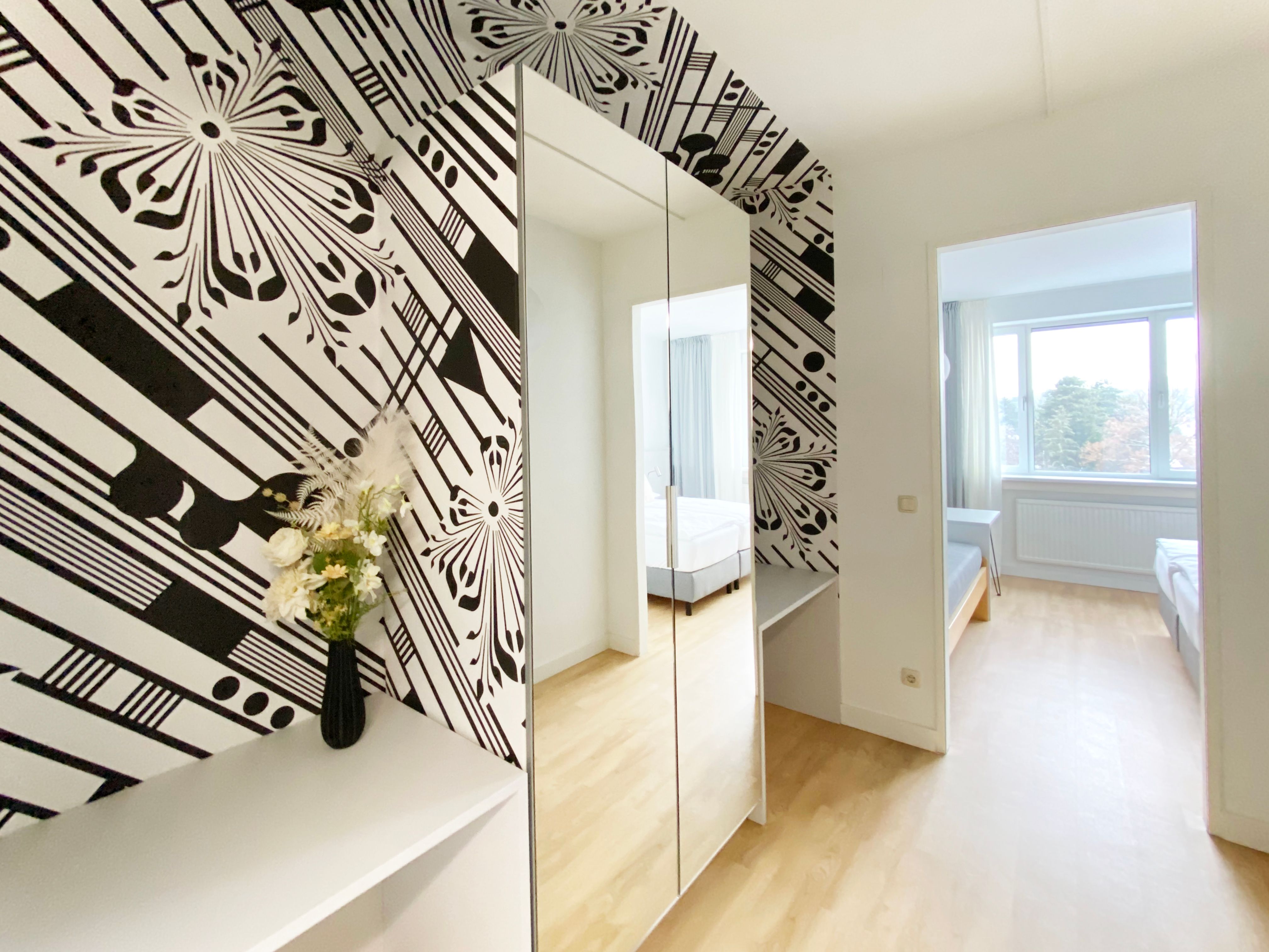 Modern guest room with black and white wallpaper, mirror, flowers and a view of a bed through a door.