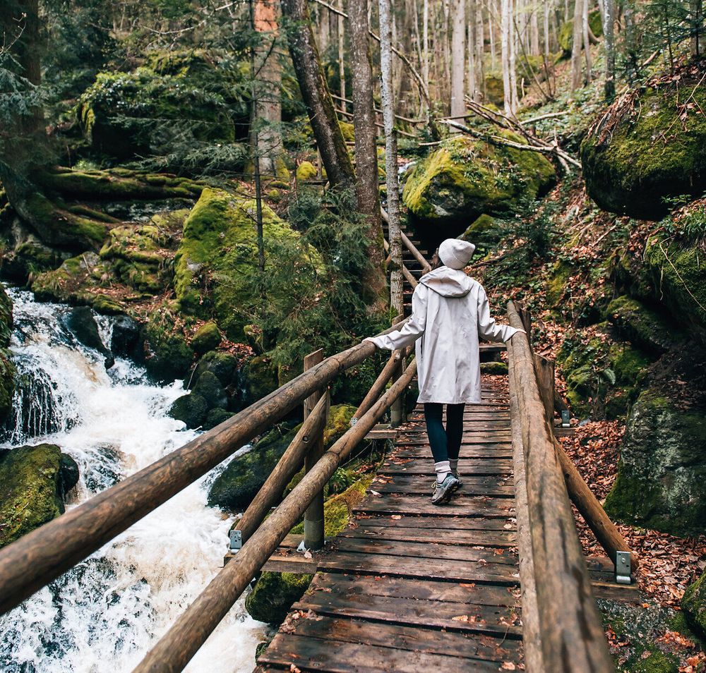 Ein sanfter Wasserfall plätschert über die moosbedeckten Steine, während Wanderer auf der Holzbrücke die Ruhe der Natur genießen. Umgeben von hohen Bäumen und frischem Grün, lädt dieser Ort dazu ein, die Seele baumeln zu lassen und die Schönheit der Umgebung zu erkunden.